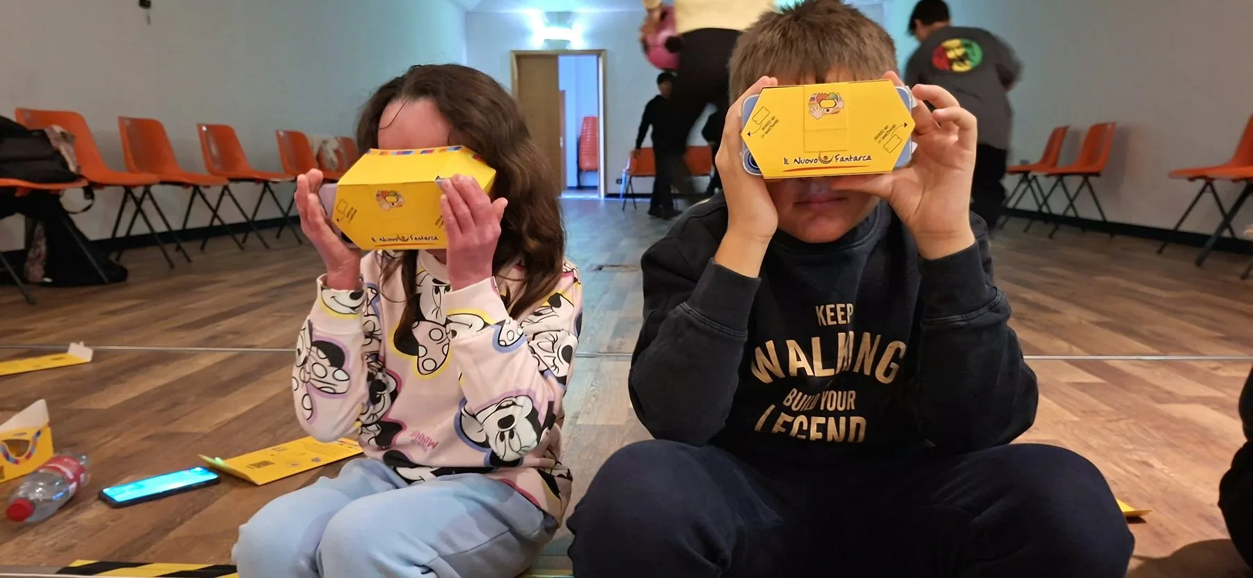 Due bambini seduti a terra tengono davanti agli occhi i visori di cartone gialli di Cinema in Ospedale